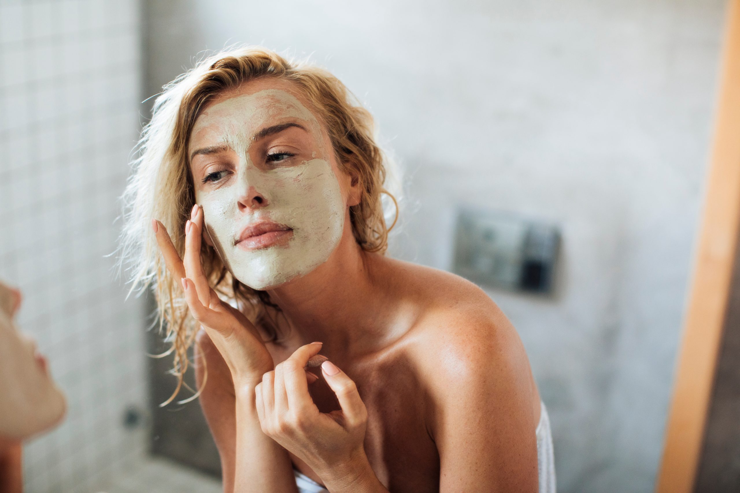 Beautiful blonde Caucasian woman standing in her bathroom and putting cosmetic mask on her face.